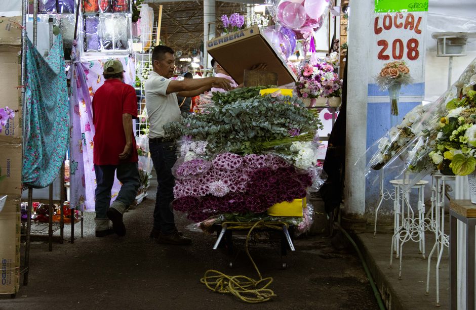 A florist at a flower stall arranging a bouquet of fresh pink and purple chrysanthemums, with various floral arrangements and bouquets displayed on tables and shelves around them. The florist is dress