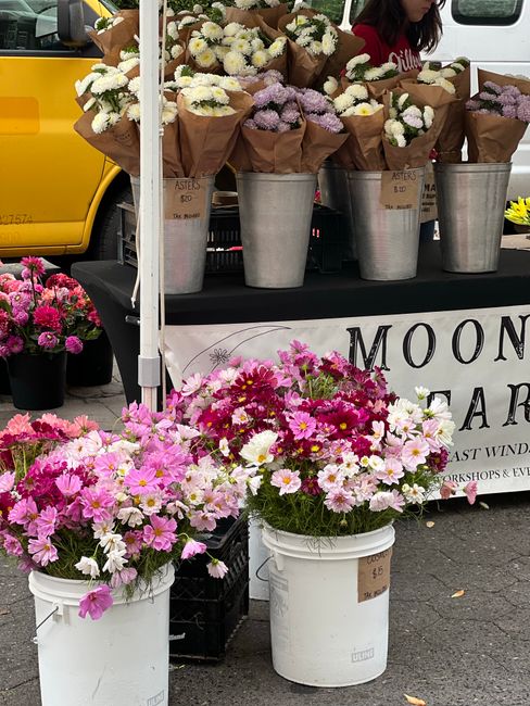 A display of fresh flower bouquets arranged in brown paper wrappers, placed in large metallic buckets on a black table at a flower stall. The bouquets feature white, lavender, and light pink flowers, 