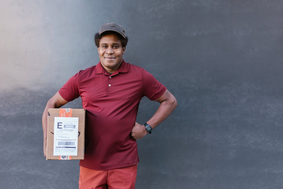 A smiling delivery person with dark skin and short curly hair, wearing a maroon polo shirt, matching cap, and a watch on their left wrist, stands against a plain dark grey wall. They are holding a sma