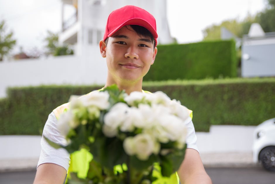 A delivery person wearing a bright red cap and a high-visibility vest is standing outdoors, holding out a bouquet of fresh white roses and green foliage towards the camera. The bouquet is wrapped in c