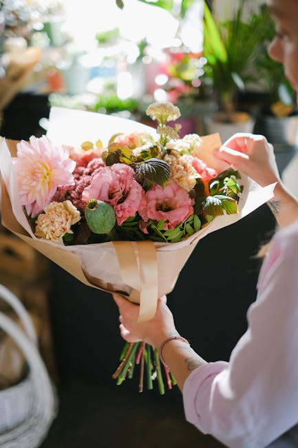 A person holding a large bouquet of fresh flowers, including pink dahlias, peonies, and white carnations, wrapped in cream and brown paper with green foliage accents. The bouquet is arranged in a lush