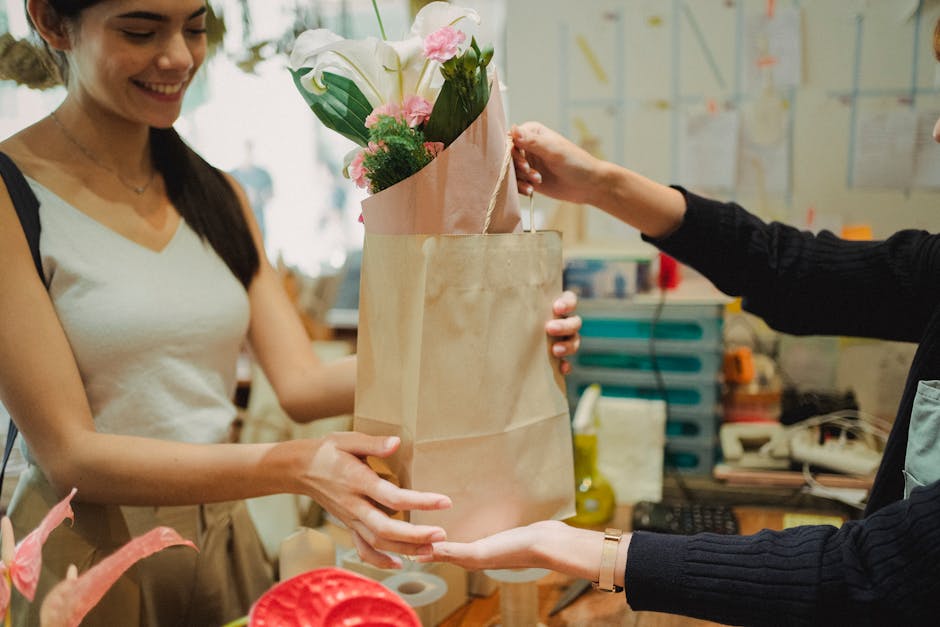 A female florist in a sleeveless beige top is smiling while handing over a bouquet arranged with white lilies, pink roses, and green foliage to a customer. The bouquet is wrapped in light pink paper a