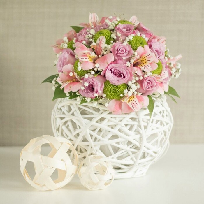 At the heart of a serene Erith interior, this floral table arrangement commands attention with its exquisite mix of colour, texture, and gentle grace. The canopy of the bouquet is composed of pastel pink and lavender roses, their soft layers unfolding amid richly marked pink alstroemeria. The unique forms of lime green button chrysanthemums offer lively pops of brightness, while wisps of cloudlike white baby's breath (Gypsophila) weave between blooms, lending a subtle, airy softness. Shining dark green leaves peep out, supplying a lush underpinning and organic contrast. Every stem is expertly situated within a striking white vase, its open basket-weave reminiscent of the artisanal crafts found in Erith's creative quarters. The vase's substantial, rounded body grounds the bouquet, while two beautifully crafted spheres-one larger than the other, both made from pale woven material-rest at its side on the immaculate tabletop. Their natural rattan-like structure provides a visual echo to the vase's intricate design, further enhancing the arrangement's tactile richness. The backdrop is a quiet, softly textured beige, allowing the joyful tones to take center stage. Perfect as a centerpiece for local wedding tables, special event gatherings in Erith, or to bring a breath of celebration and thoughtful design into one's home, this image embodies freshness, craftsmanship, and the welcoming heart of Erith's floral community.