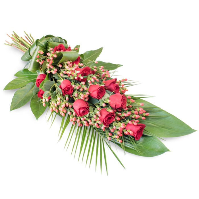 An elegant, teardrop-style funeral spray of red roses and berries is presented against a pristine white background, its diagonal placement creating a sense of gentle, forward movement. This arrangement, similar to those often chosen for services in Erith's churches and small chapels, combines classic flower choices with a refined, contemporary structure. The central focus is a continuous line of red roses arranged along the length of the spray. Their colours range from deep, velvety crimson to slightly lighter ruby tones, and their stages of opening vary-some tightly furled, others partially open, with layers of petals forming delicate spirals. Interlaced with these roses are clusters of pinkish-red Hypericum berries, each berry small, round, and smooth, clustered together like tiny jewels. These berries soften the visual weight of the roses and add a gentle, almost playful texture without breaking the solemn mood. Beneath this floral line stretches a carefully constructed base of foliage. Broad, dark green leaves with a glossy finish provide structure, echoing the long, tapered form of the spray. From the lower right, fine palm fronds fan outward in graceful arcs, extending the silhouette and bringing a flowing, feathery element that contrasts with the solid, rounded rose heads. Additional slender stems and sprigs of greenery weave between the flowers, filling gaps and creating a rich, full appearance, as if the piece had been assembled from freshly cut stems just delivered to a florist near Erith town centre. At the upper left, the stems are bound tightly and wrapped with a smooth band of green leaf, giving a polished, handcrafted finish. Overall, the spray feels dignified and serene, a fitting floral expression of love, remembrance, and respect for a final farewell in Erith.